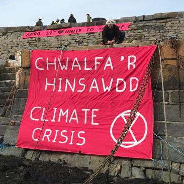 Person dropping banner over edge of sea wall that says "Climate Crisis" in Welsh & English